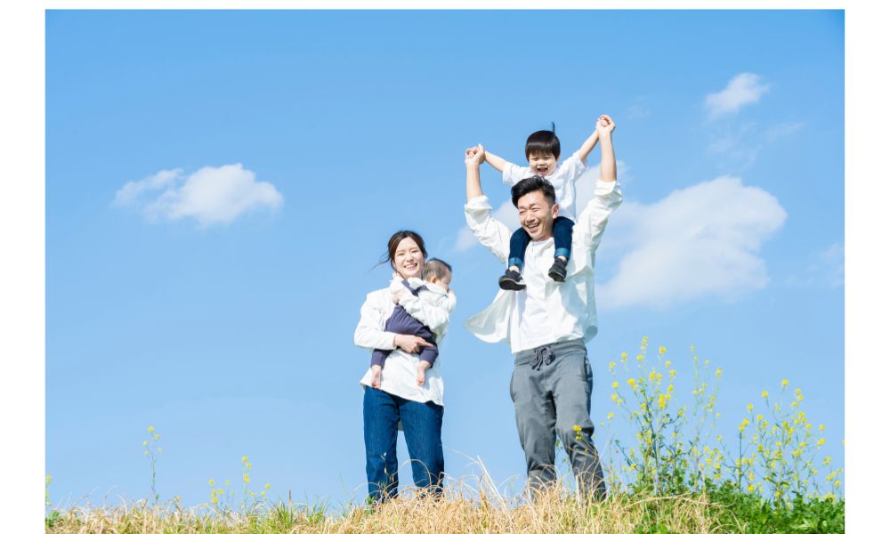 青空の下、父親が子どもを肩車し、母親がもう一人の子を抱っこして並ぶ家族