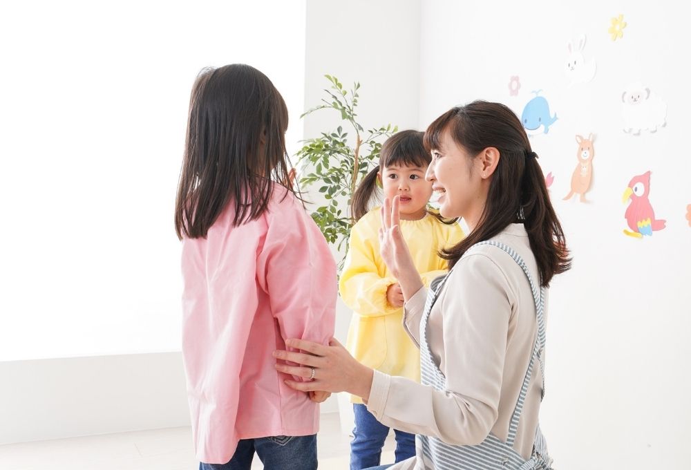 保育士が2人の幼児と笑顔で向き合う保育室の場面