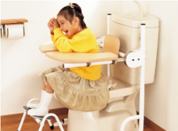 ハートリーフレスト　テーブルタイプ.
黄色い服の女の子が補助テーブル付きのトイレチェアに座り、楽しそうに笑っている。