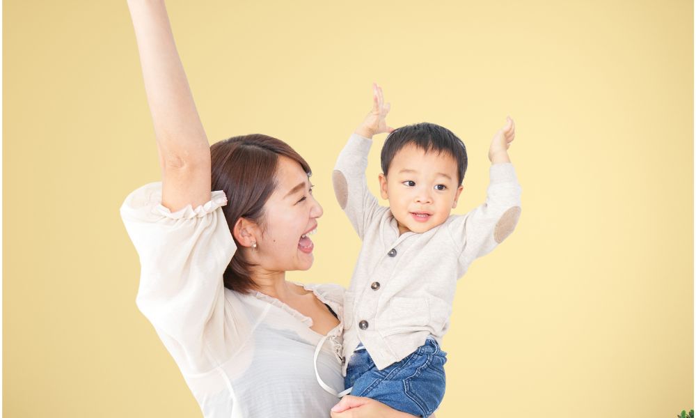 黄色い背景の前で、トイトレ成功を喜び両手をあげて笑う母親と子ども。