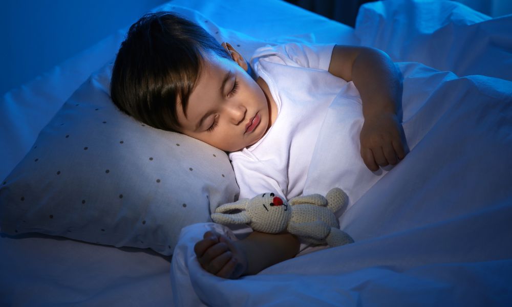 い青い照明の中、白いシャツを着た男の子がぬいぐるみを抱いて寝ている