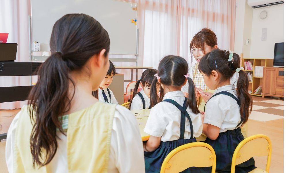 保育園で数人の子どもたちが机を囲み、先生が優しく声をかけながら活動している様子