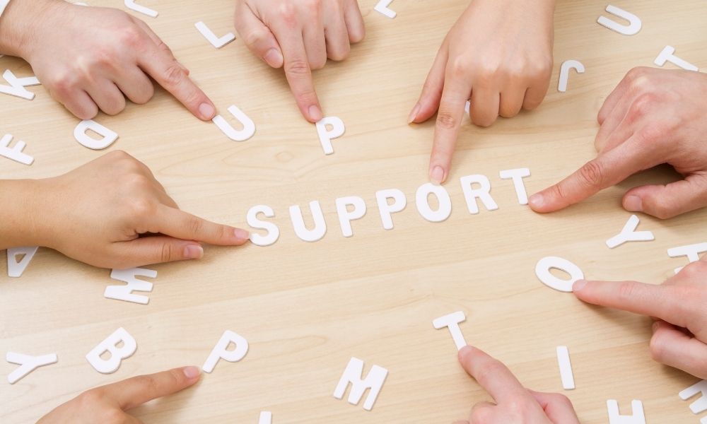 複数の人の手が机の上のアルファベット文字を指し示し、「SUPPORT(支援)」という言葉を形づくっている様子。協力し合いながら支え合う関係性や、家族・周囲のサポート体制を象徴するイメージ。