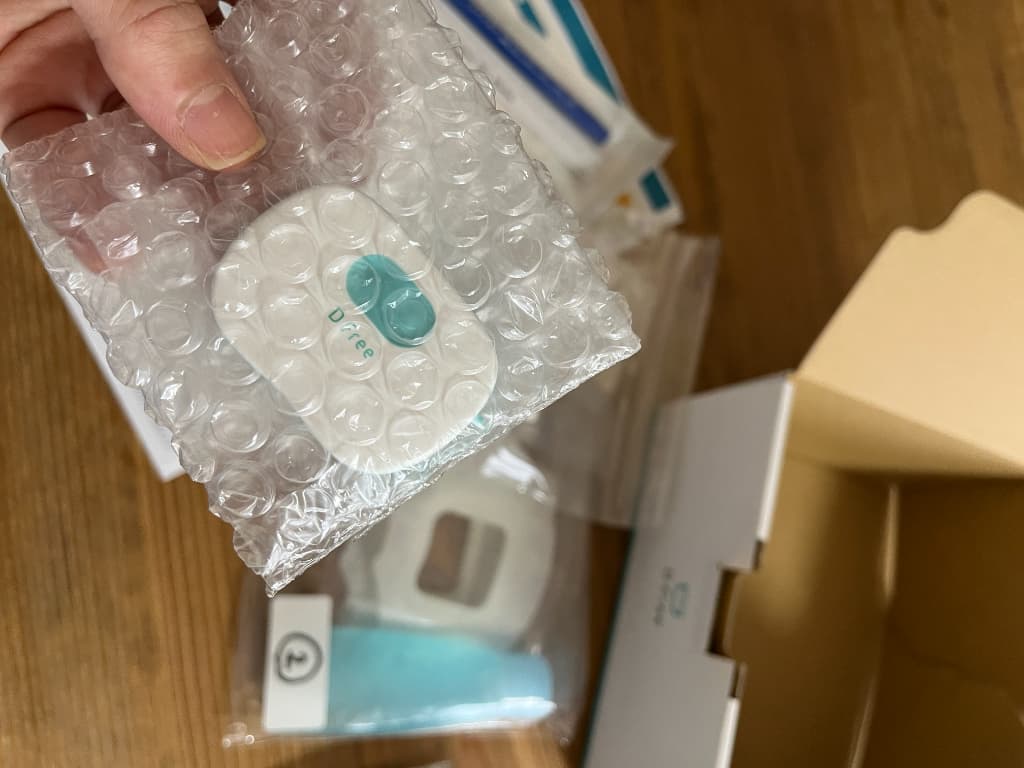 緩衝材に包まれたDFree本体を手に持ち、周囲に箱や付属品が並ぶ開封直後の様子。トライアル開始時に届く内容物の一部が分かる写真。