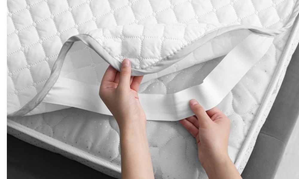 キルティング加工されたマットレスプロテクターを、手でめくって装着部分を確認している様子です。ゴムバンド付きで、マットレスにしっかり固定できる構造が分かります。