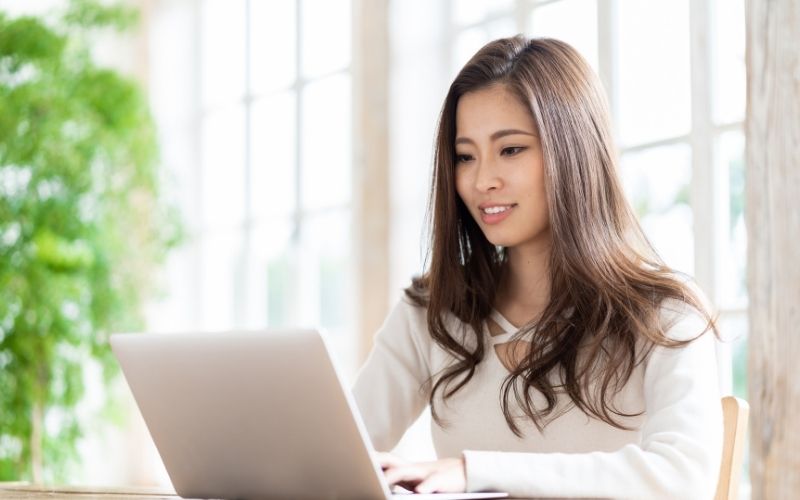 明るい室内で、ノートパソコンを操作している女性の様子が写っています。窓からの自然光が入り、落ち着いた雰囲気の中で作業している場面です。