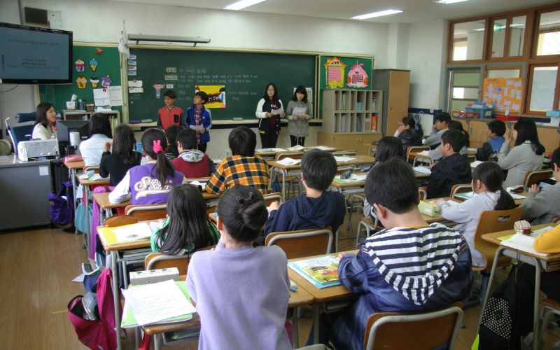 教室の後方から全体が見えている様子。生徒が着席し、授業を受けている。黒板の前には数人の生徒が立っており発表している様子