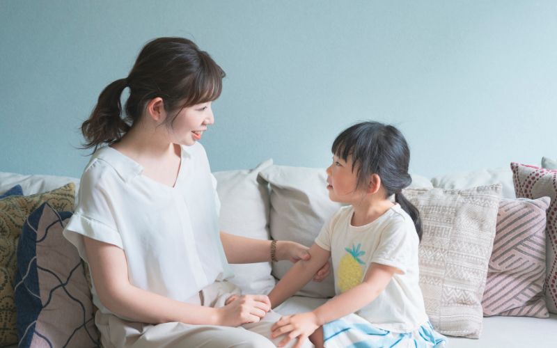 ソファに座った母親と小さな女の子が向かい合って話している様子。母親がやさしく腕に手を添え、子どもも見上げながら安心した表情でやり取りをしている