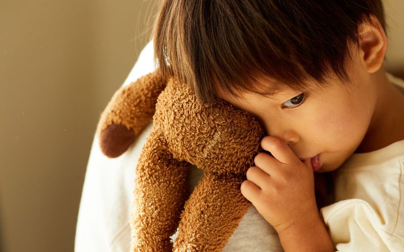 小さな子どもが茶色のぬいぐるみをぎゅっと抱きしめ、少し不安そうな表情で寄り添っている様子。やわらかな光の中で、安心を求めるような雰囲気が感じられる