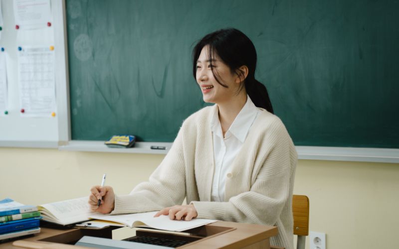 教室の机に座り、ノートを取りながら微笑む教師の様子。黒板を背景に、落ち着いた雰囲気の中で授業や指導を行っている場面を写した写真