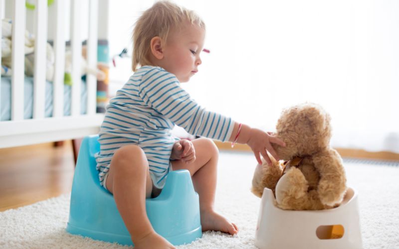 室内でおまるに座る幼児が、そばに置かれたぬいぐるみに手を伸ばして遊んでいる様子。トイレトレーニング中に、遊びを取り入れてリラックスしている場面を写した写真