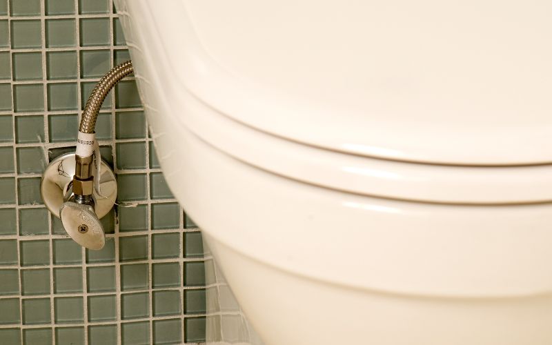 トイレの側面と給水管の止水栓部分をクローズアップした写真。緑色のタイル壁を背景に、金属製のホースとバルブが取り付けられている様子が写っている