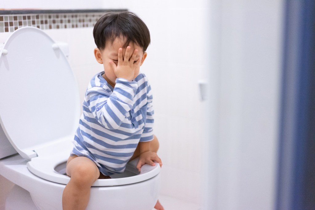 洋式トイレに座った小さな男の子が、手で顔をおおって困ったような様子をしている。トイレトレーニング中の場面をイメージさせる写真