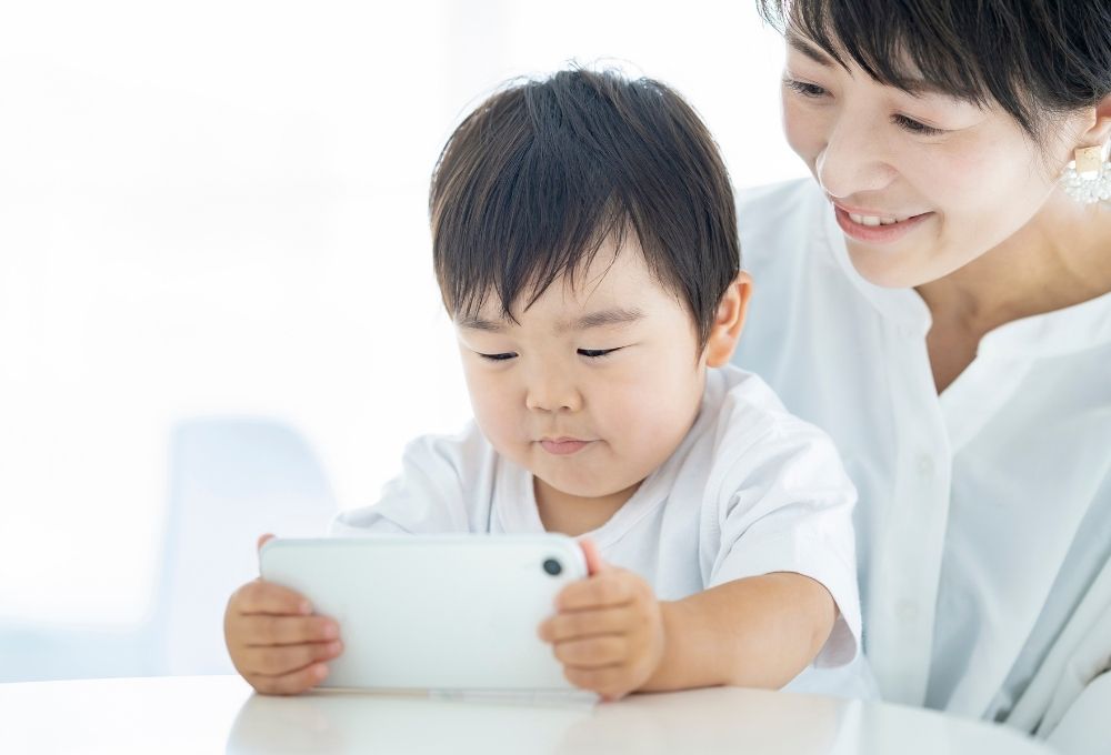 テーブルの前で、小さな子どもがスマートフォンまたは小型タブレットを両手で持って画面を見ている。隣で母親が笑顔で見守っており、親子で一緒に画面を見ている様子の写真