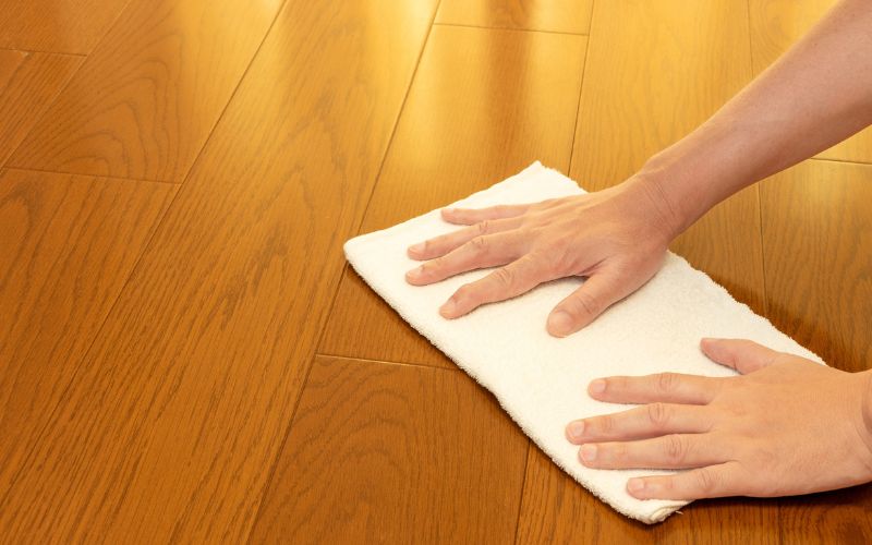 木のフローリングの床を、白い布で手拭き掃除している様子。両手で布を押さえながら床を拭いており、室内の掃除や片付けの場面を表した写真