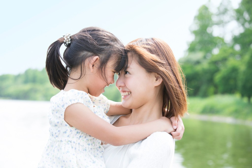 屋外の自然の中で、母親が女の子を抱き上げて向かい合い、額を寄せながら笑い合っている様子。背景には水辺と緑の木々が広がり、明るくあたたかい雰囲気の写真