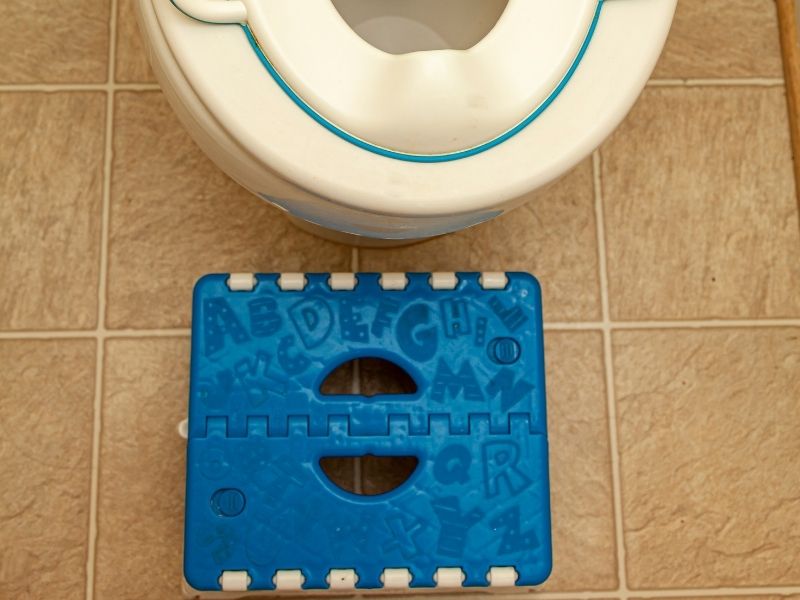 子ども用の補助便座が置かれたトイレの前に、青い折りたたみ式の踏み台が設置されている様子を上から撮影した写真。踏み台にはアルファベットの模様があり、子どもがトイレに座るときに足を乗せるための補助用品