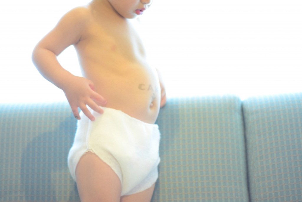 白い紙おむつを履いた幼い子どもがソファの上に立ち、自分のお腹まわりを見ながら手で触れている様子。背景は明るく、淡い色のソファが写っている