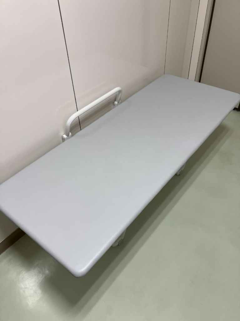 白い壁に取り付けられた手すりの下に、大きなグレーのクッション台が設置されている。床は淡い緑色で、バリアフリートイレや多目的トイレ内にある介助用のオムツ交換・着替え用ベッドのように見える。