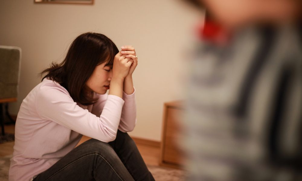 室内で女性が膝を抱え、手を組んで顔を伏せている様子。疲れや悩みを抱えているような表情で、介護や育児の負担を感じている場面。