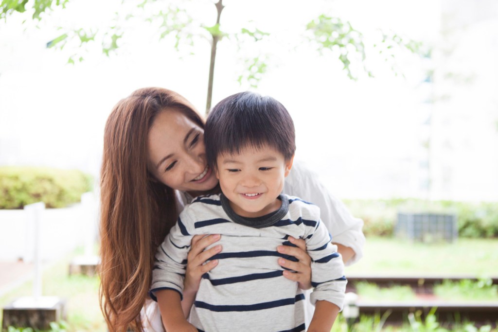 屋外で母親が小さな子どもを後ろから抱きしめ、二人とも笑顔を見せている様子。安心感と親子の温かい関わりを感じる場面。