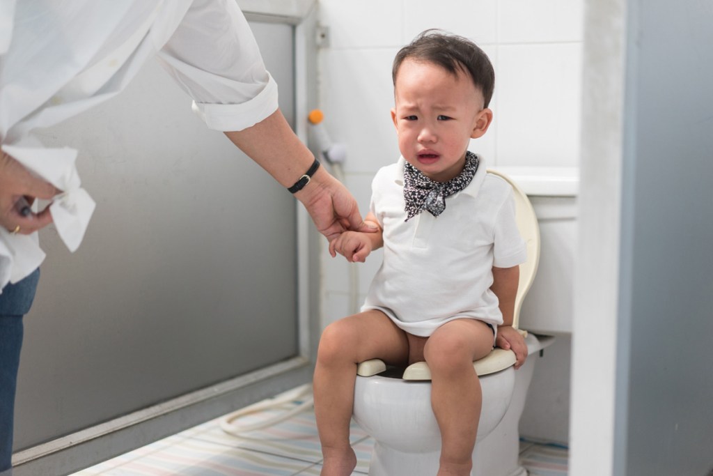トイレに座っている幼い子どもが不安そうな表情をしており、大人が手を握ってサポートしている様子。トイレトレーニング中の戸惑いや難しさを表している。