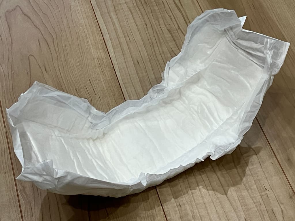 木目の床の上に広げられた使い捨てのおむつの写真。白色で吸収パッド部分が見え、成人用サイズの紙おむつの形状をしている