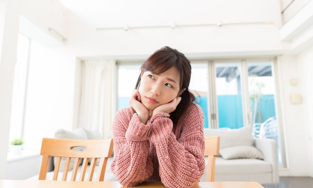 A young woman sitting at a dining table with a thoughtful expression, resting her chin on her hands, in a bright and airy room.