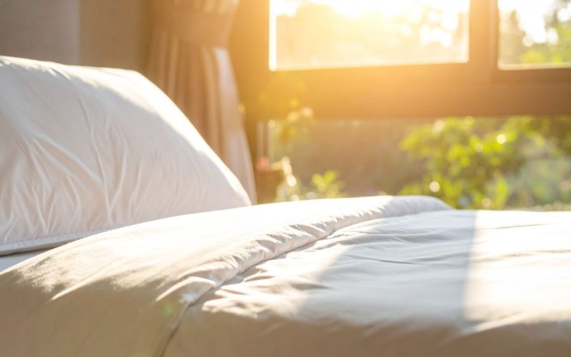 A close-up view of a neatly made bed with white sheets and a pillow, illuminated by warm sunlight streaming in through a window.