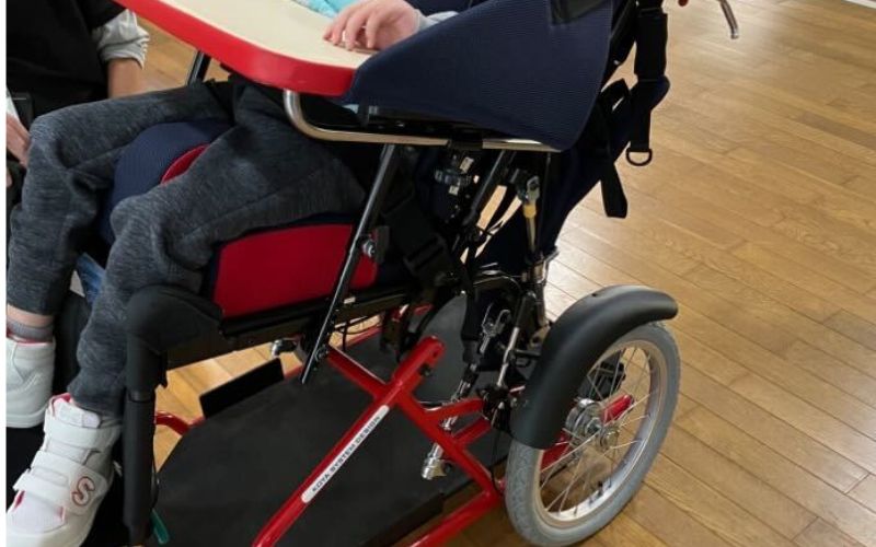 車椅子に座った子どもの様子を写した写真。姿勢保持機能付きの椅子やテーブルが取り付けられており、身体的サポートや日常生活支援、排泄支援の環境をイメージさせる構図。
