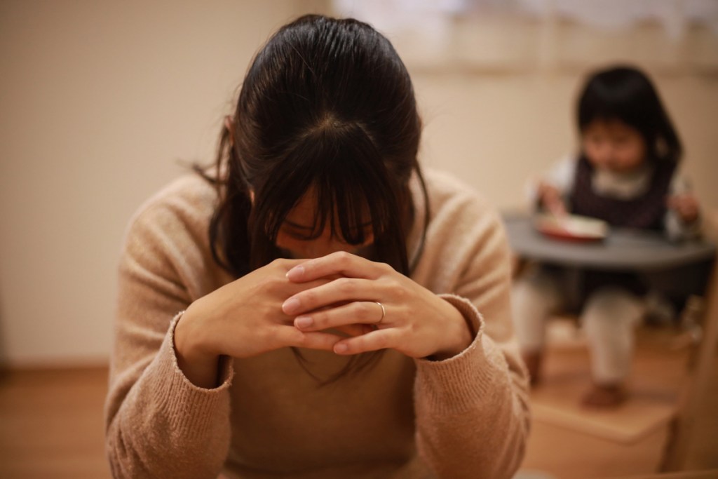 室内で、女性が両手を組んでうつむき、不安そうな様子を見せている。背景には子ども用チェアに座った子どもがぼんやり写っており、育児の悩みやストレスをイメージさせる写真。