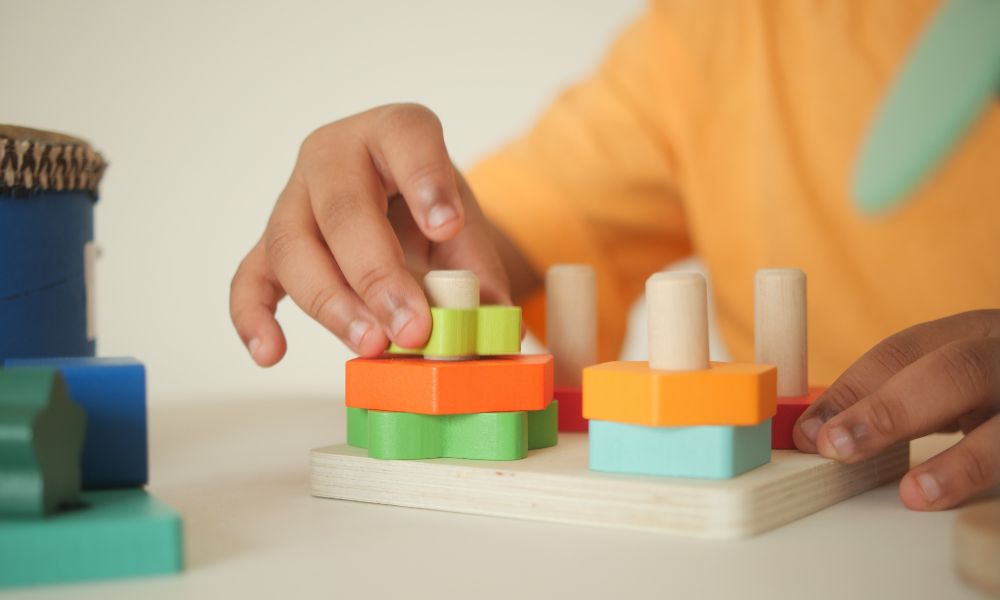 子どもがカラフルな木製の知育玩具で遊んでいる場面のクローズアップ。手で形のついたブロックを棒にはめようとしており、机の上に複数の色鮮やかなパーツが並んでいる。