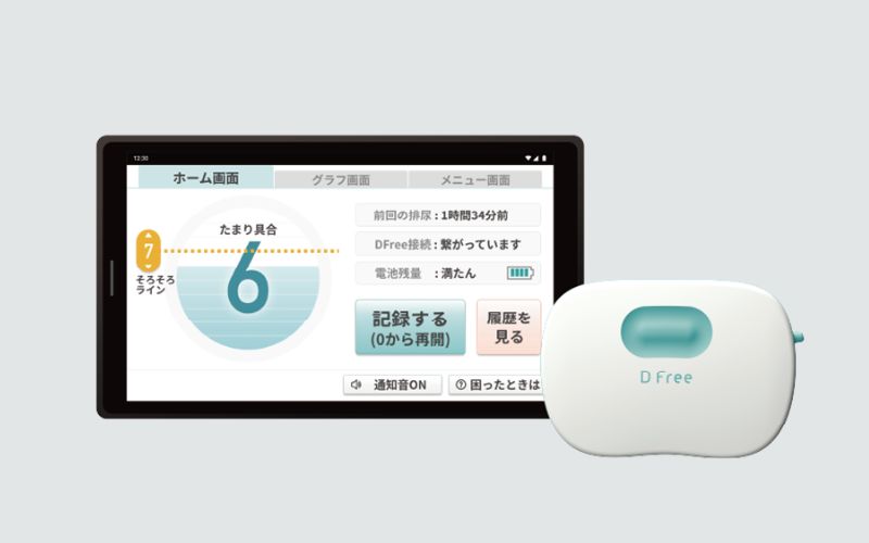 タブレット画面と小型デバイスが並んだ画像。画面にはおむつの状態管理アプリのような表示があり、「たまり具合」や排尿時刻、履歴確認などの情報が表示されている。右側には白色のセンサー機器が置かれている。