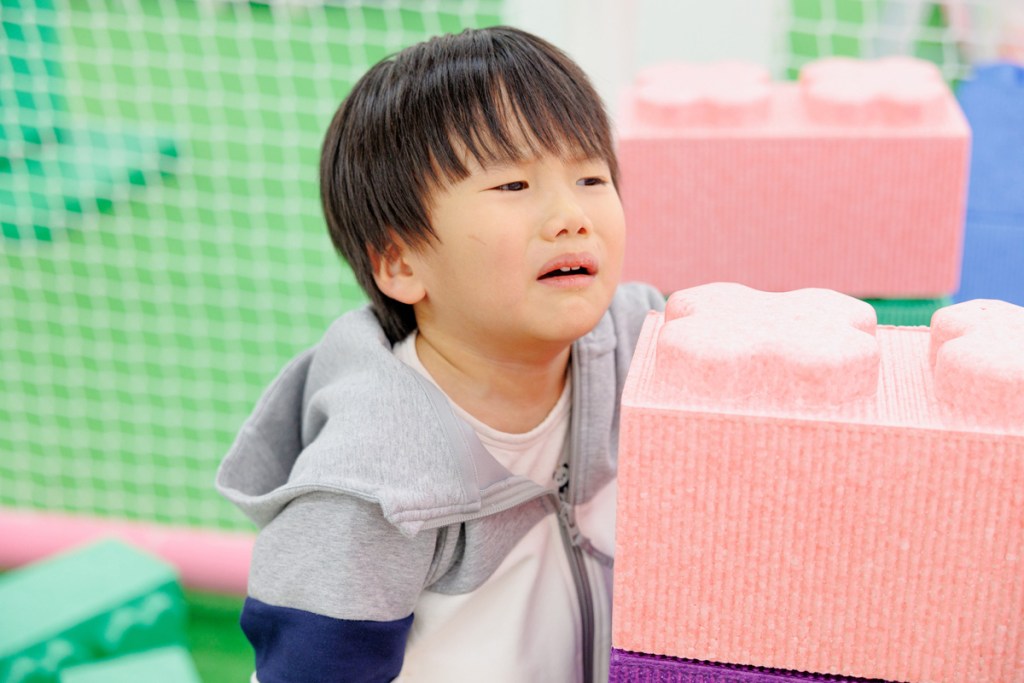 屋内の遊び場のような場所で、ピンク色の大きなブロックのそばにいる子どもの様子。涙ぐんだような表情で、困ったり不快そうに見える。背景には緑色のネットや遊具が写っている。