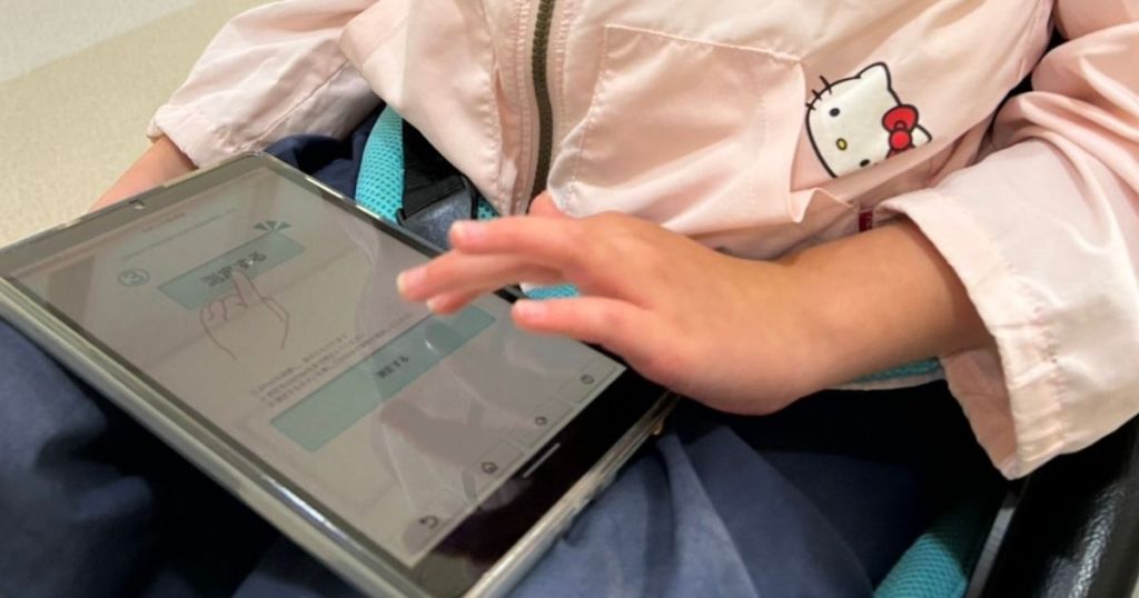 車椅子に座る子どもがタブレットを操作している様子。排尿タイミングを支援するDFreeアプリを使用している場面。